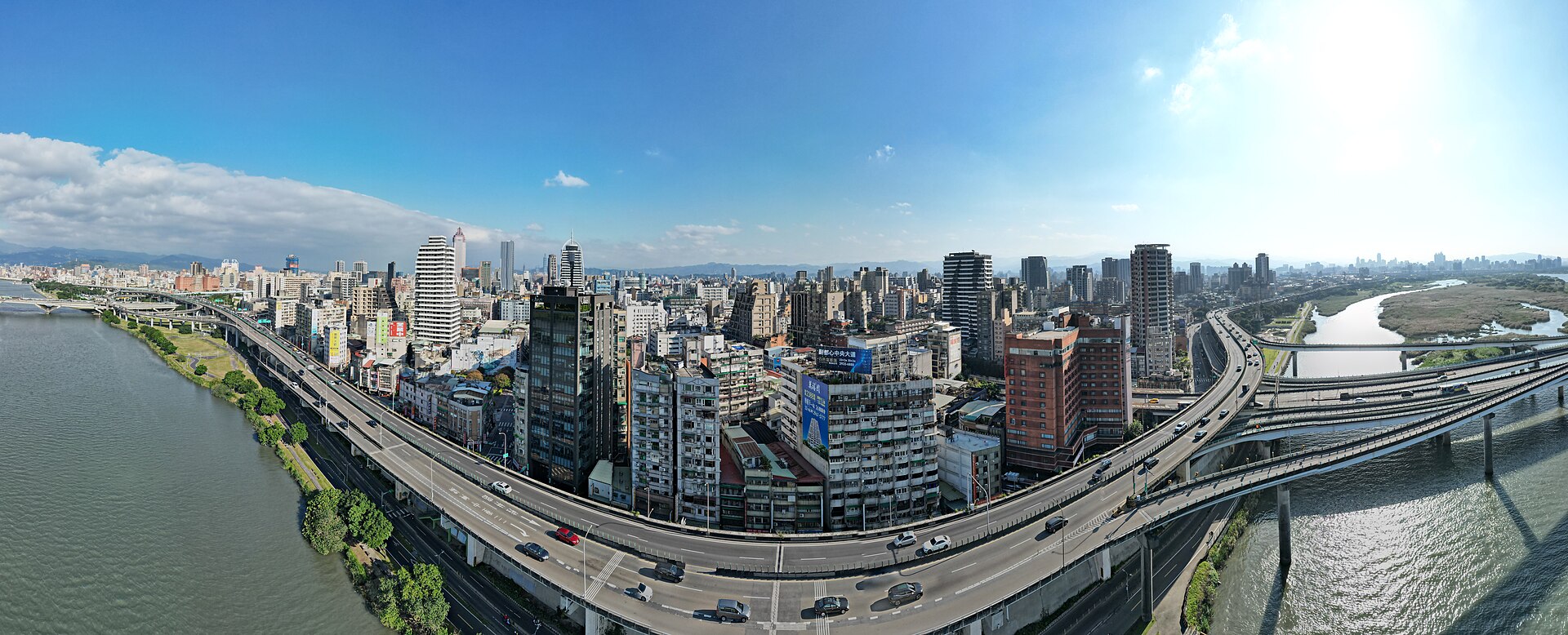 從淡水河上空俯瞰台北市西區,高架快速道路沿著河岸穿過密集的市區大樓,遠方是丘陵與市中心天際線