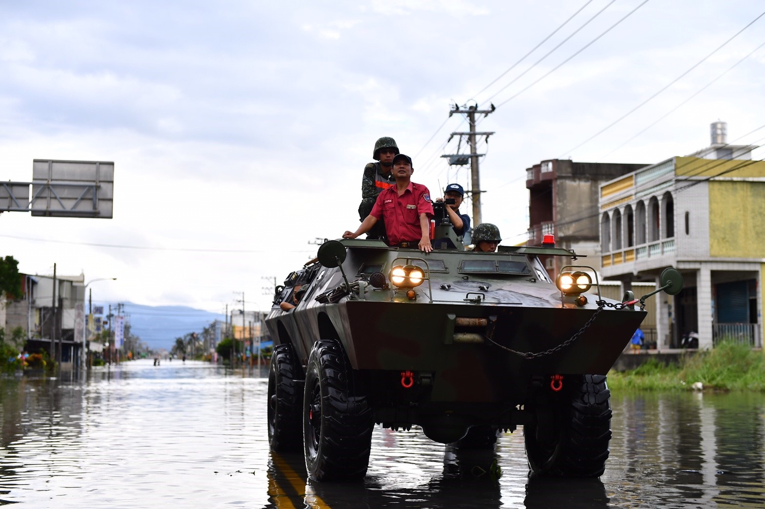 國軍裝甲車行駛在屏東淹水的街道上,車頂站著士兵與一名穿紅色制服的消防人員,兩旁街屋泡在水中